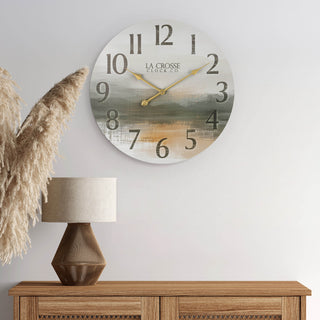 Lifestyle image of the 404-3461 24-inch Daybreak Wall Clock shown hanging on a white wall above a rattan and wood cabinet with a lamp. The clock features an open face design of an impressionistic sunrise in muted tones, distressed numbers, and gold metal hands.