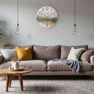 Lifestyle image of the 404-3461 24-inch Daybreak Wall Clock shown hanging on a grey wall above a taupe couch with neutral pillows. The clock features an open face design of an impressionistic sunrise in muted tones, distressed numbers, and gold metal hands.