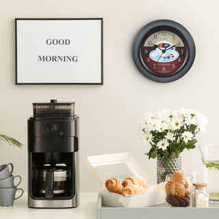 Lifestyle image of the 404-3623C 9-inch Cafe Wall Clock hanging on a wall above a coffee bar with a "Good Morning" sign. The clock features a coffee motif dial, a thick black frame, and black metal hour and minute hands.