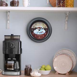 Lifestyle image of the 404-3623C 9-inch Cafe Wall Clock hanging on a wall above a counter with a coffee pot, coffee, apples, sugar cubes, and plates. The clock features a coffee motif dial, a thick black frame, and black metal hour and minute hands.