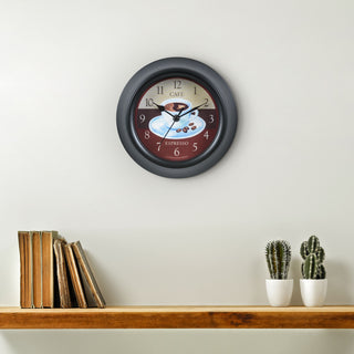Lifestyle image of the 404-3623C 9-inch Cafe Wall Clock hanging on a wall above a wood shelf with books and a cactus. The clock features a coffee motif dial, a thick black frame, and black metal hour and minute hands.