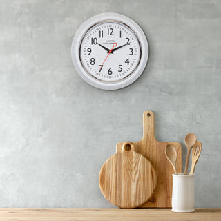 Lifestyle image of the 404-3624W 9.5-inch Diner Clock – White, shown hanging on a textured grey wall above a wood counter with white and wood accents. The clock features a retro-style white frame with a white dial, chrome bezel, black numbers and minute markings, black metal hands, and a red second hand.
