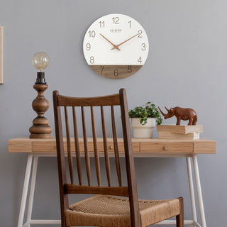 Lifestyle image of the 404-3630B 12-inch Sierra Wood Wall - White, hanging on a gray wall above a Scandinavian style desk of white and light wood with a lamp, a potted plant, a wood rhinocerous sculpture and a Transitional style wood chair. The clock features laser-cut numbers, a black and wood open-face dial, and wood hour and minute hands.
