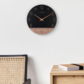 Lifestyle image of the 404-3630C 12-inch Sierra Wood Wall Clock - Black, the clock is shown hanging on a wall above a side table and a wicker chair. The clock features a black and wood split design open-face dial, with laser cut numbers, and wood hour and minute hands.