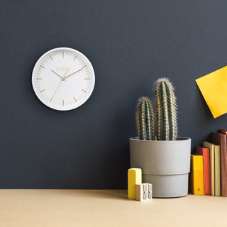 404-3715 A minimalist white round wall clock with gold hands and gold hour markers hangs on a dark charcoal wall above a beige tabletop. The scene includes a potted cactus in a gray pot, stacked books, yellow blocks, and a yellow paper sheet, creating a styled interior setting.