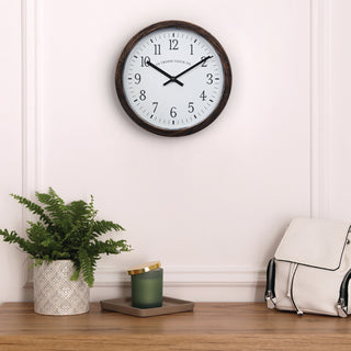 404-3825HC A round wall clock with a dark wood frame and white face hangs on a white paneled wall above a wooden surface. A potted green fern, a green candle in a tray, and a cream backpack are arranged below.