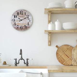 Lifestyle image of the 404-3828K 11-inch Whitman Wall Clock shown hanging on a cream wall in a farmhouse style white kitchen over the sink. The clock features an open-faced dial with a weathered wood design, black numbers, and black metal hands.