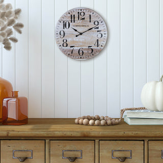 Lifestyle image of the 404-3828K 11-inch Whitman Wall Clock shown hanging on a cream shiplap wall over a rustic side table with fall decorations. The clock features an open-faced dial with a weathered wood design, black numbers, and black metal hands.