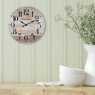 Lifestyle image of the 404-3828K 11-inch Whitman Wall Clock hanging on a sage green wood shiplap style wall over a rustic wood buffet table with white dishes. The clock features an open-faced dial with a weathered wood design, black numbers, and black metal hands.