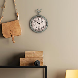 Lifestyle image of the 9-inch 404-3828 Pocket Watch Wall Clock hanging on a tan wall above a metal entry table with rustic boxes and a ball of thread. The clock features a green patina pocket-watch-style frame with a parchment dial, black numbers, and black stylized hands.