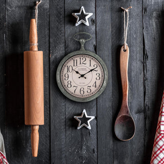 Lifestyle image of the 404-3828 9-inch Pocket Watch Wall Clock hanging on a black-stained rustic wood wall with a wood rolling pin, a wooden spoon, star-shaped cookie cutters, and a red checked flour sack towel. The clock features a green patina pocket-watch-style frame with a parchment dial, black numbers, and black stylized hands.