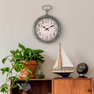 Lifestyle image of the 404-3828 9-inch Pocket Watch Wall Clock hanging above a wood cabinet with a model sailboat, a nautical lamp, and a plant. The clock features a green patina pocket-watch-style frame with a parchment dial, black numbers, and black stylized hands.