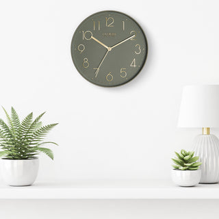 Lifestyle image of the 404-3831G 12-inch Paxton Wall Clock - Green, with Silent Sweep, hanging on a white wall above a white shelf with a white lamp and plants in white vases. The clock features a flush-mount green frame, a matching color dial, brass raised numbers, and brass-finished hour, minute, and second hands.