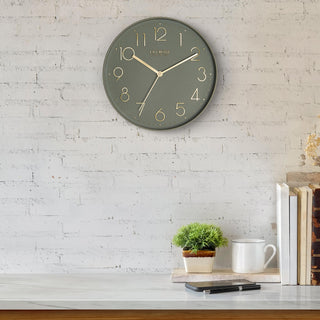 Lifestyle image of the 404-3831G 12-inch Paxton Wall Clock - Green, with Silent Sweep, hanging on a white-washed brick wall above a marble countertop with books, a plant, and a phone. The clock features a flush-mount green frame, a matching color dial, brass raised numbers, and brass-finished hour, minute, and second hands.