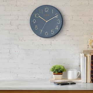 Lifestyle image of the 404-3831SP 12-inch Paxton Wall Clock - Speckled, with Silent Sweep, hanging on a white-washed brick wall above a marble countertop with books, a plant, and a phone. The clock features a flush-mount speckled blue frame, a matching color dial, brass raised numbers, and brass-finished hour, minute, and second hands.