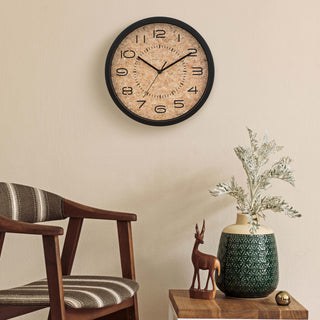 Lifestyle image of the 404-3832 12-inch Felix Cork Wall Clock shown hanging on a tan wall above a mid-century modern wood chair with striped neutral fabric and a side table with a wood deer sculpture and a forest green vase. The clock features a cork dial with laser-cut numbers and time markings in a black case and hour, minute, and second hands.
