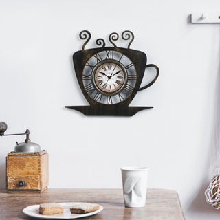 404-3833B A wall-mounted coffee cup-shaped clock with a dark wood finish and Roman numeral face, shown above a table with a cup and bread in a styled interior setting.