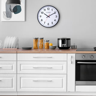 Lifestyle image of the 404-3841 16-inch Commercial Wall Clock shown hanging on a grey wall in a modern kitchen with white cabinets. The clock features bold black numerals, black hour markings, a white dial, black hour and minute hands, and a red second hand in a black flush-mount frame.