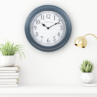Blue wall clock on a white wall with decorative elements like books, plants, and a lamp.