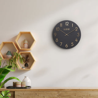 Black wall clock on a white wall with wooden shelves and plants in the background