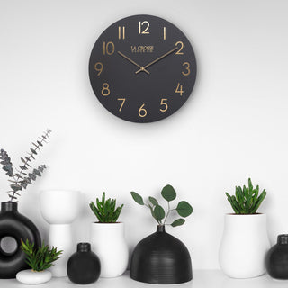 404-3841C Black wall clock with gold numbers on a white wall, surrounded by decorative vases and plants.