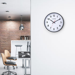 Lifestyle image of the 404-3841 16-inch Commercial Wall Clock shown hanging on a white wall in an industrial inspired loft office building. The clock features bold black numerals, black hour markings, a white dial, black hour and minute hands, and a red second hand in a black flush-mount frame.