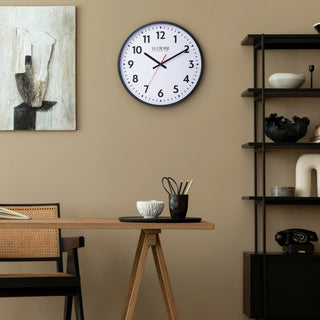 Lifestyle image of the 404-3841 16-inch Commercial Wall Clock shown hanging on a dark golden-tan wall next to a black book shelf above a wood desk in a transitional decor space. The clock features bold black numerals, black hour markings, a white dial, black hour and minute hands, and a red second hand in a black flush-mount frame.