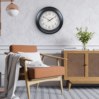 A round wall clock with a black frame and white face hangs on a textured white wall above a mid-century modern armchair with an orange-brown seat cushion and wooden frame. A light wood cabinet with a cane-style front is to the right, and a hanging pendant light is visible on the left.