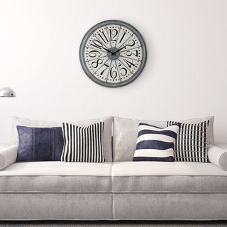 A round wall clock with a gray frame and black-and-white numerals hangs above a light gray sofa with navy and striped throw pillows in a bright living room.