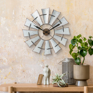 A rustic metal windmill-style wall clock with distressed silver blades and black hands, mounted on a textured beige wall above a wooden console table styled with a potted plant, small figurines, and books.