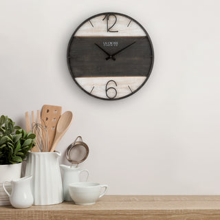 A round wall clock with a rustic three-stripe design in black, dark gray, and whitewashed wood, hanging on a white wall above a kitchen counter with white ceramic mugs and utensils.