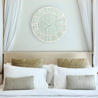 A neutral decorated bed with tan, gray, and white pillows with the 404-4060 23.5‑inch Harbor Tides wall clock hanging above the light wood frame bed. The clock features a distressed light teal wooden frame with an open face dial design, wood cut out Roman numerals in a light distressed white, and decorative metal hour and minute hands.