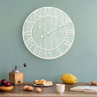 Lifestyle image of the 404-4060 23.5‑inch Harbor Tides wall clock features a distressed light teal wooden frame with an open face dial design, wood cut out Roman numerals in a light distressed white, and decorative metal hour and minute hands. The clock is featured hanging on a light seaglass green wall above a rustic table with eggs, squash, book, coffee cup, and a vintage coffee grinder.