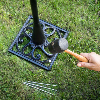 Lifestyle image of the 425-34188 Outdoor Dual-Sided Metal Pedestal Clock showing a hand holding a rubber mallet installing the wrought iron base by staking it into the grass with the included metal stakes.