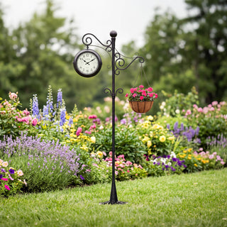 Lifestyle image of the 425-34188 Outdoor Dual-Sided Metal Pedestal Clock shown staked in a grassy lawn with a hanging flower basket in front of a flower bed. The clock features a temperature dial in a Fahrenheit scale, a humidity dial, decorative black metal hands, black numbers on a flecked white dial surrounded by a dark brown metal frame., with a 72-inch decorative scrollwork pedestal with a plant hanger.