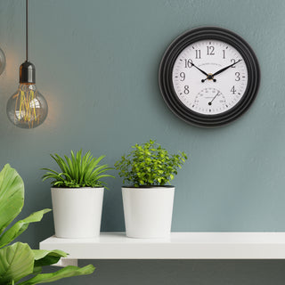 Lifestyle image of the 433-3020B 8-inch Colby Indoor/Outdoor Wall Clock with temperature hanging on a blue wall above a white shelf with white vases with plants. The clock features a dark decorative frame, a white dial with black numbers, a Fahrenheit temperature gauge, and black metal hands.
