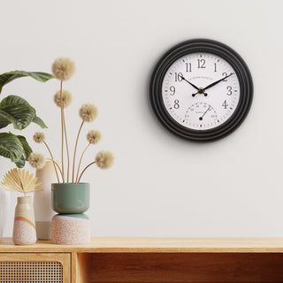 Lifestyle image of the 433-3020B 8-inch Colby Indoor/Outdoor Wall Clock with temperature hanging on a cream wall over a Mid-Century Modern side table with vases. The clock features a dark decorative frame, a white dial with black numbers, a Fahrenheit temperature gauge, and black metal hands.