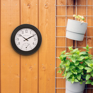 Lifestyle image of the 433-3020B 8-inch Colby Indoor/Outdoor Wall Clock with temperature hanging on a shed next to plants. The clock features a dark decorative frame, a white dial with black numbers, a Fahrenheit temperature gauge, and black metal hands.