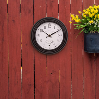 Lifestyle image of the 433-3020B 8-inch Colby Indoor/Outdoor Wall Clock with temperature hanging on a red wood fence next to a plant. The clock features a dark decorative frame, a white dial with black numbers, a Fahrenheit temperature gauge, and black metal hands.