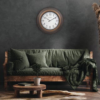 A rustic wooden wall clock with a distressed brown frame and white face hangs above a dark green textured sofa in a moody, dark room. A matching green throw blanket is draped over the sofa, and a small wooden coffee table sits in front.