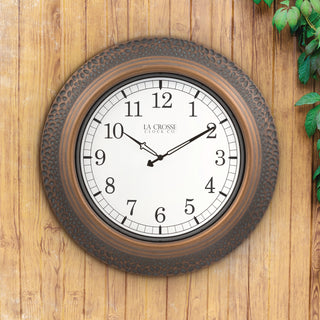 A round wall clock with a distressed dark brown textured frame and a white dial, mounted on a light brown wooden wall with green leaves visible in the top right corner. The brand 'LA CROSSE CLOCK CO.