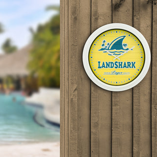 433-3841LS A circular wall clock featuring a yellow and blue Landshark logo, mounted on a brown wooden wall with a blurred resort pool background.