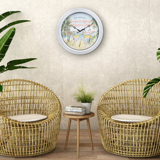 A round wall clock with a white frame and a colorful beach illustration, mounted on a textured beige wall above two woven rattan armchairs and a small wooden side table.