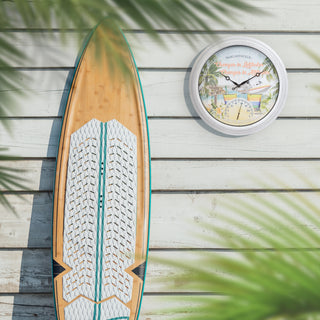 A bamboo surfboard with a white textured grip pad and teal edge details lies on a white wooden plank surface next to a round beach-themed wall clock. Palm leaf shadows are cast across the scene.