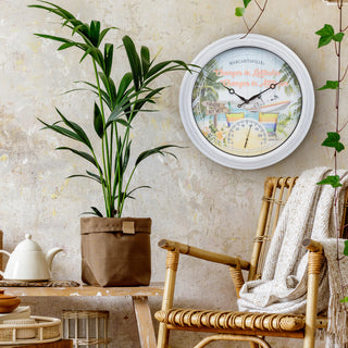 A round wall clock with a white frame hangs on a textured beige wall in a styled room. The clock face features a tropical beach scene with the text 'MARCANDISE - Change the attitude - Smiles in life - Tears in time'.