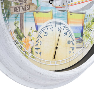 A close-up of a round wall clock with a distressed white frame and a tropical beach scene printed on the face. The clock includes a large white analog thermometer dial in the center, colorful striped umbrellas, palm leaves, flowers, and a boat illustration.