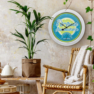 433-3841MV2T 15.75-inch Margaritaville® Indoor Outdoor Wall Clock with Temperature hangs on a tan textured wall above a rattan chair, a wood bench with a plant, and a tea service. The lime green and teal blue dial has an image of a lime with the text "it's 5' O Clock Somewhere," a temperature gauge, and black hands all surrounded by a white weathered frame.