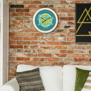 A round wall clock with a white frame hangs on a red brick wall above a white sofa with patterned and green pillows. The clock face features a turquoise and yellow design with the words 'You Can Do Somewhere.