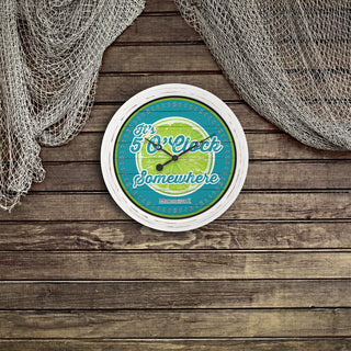 A round wall clock with a white frame and a nautical-themed face, featuring the text 'It's 5 o'clock Somewhere' over a blue ocean and green wave design. The clock is displayed on a rustic wooden plank surface with fishing nets draped in the top corners.