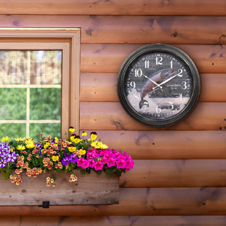 A round wall clock with a black frame and a scenic fish illustration on the face, mounted on a rustic brown wooden log wall next to a window with a colorful flower box.